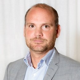 En man med kort ljusbrunt hår och vältrimmat skägg, klädd i ljusgrå kostymjacka och blåvitrandig skjorta, står framför en vit bakgrund, redo att diskutera effekterna av Lönetransparensdirektivet.