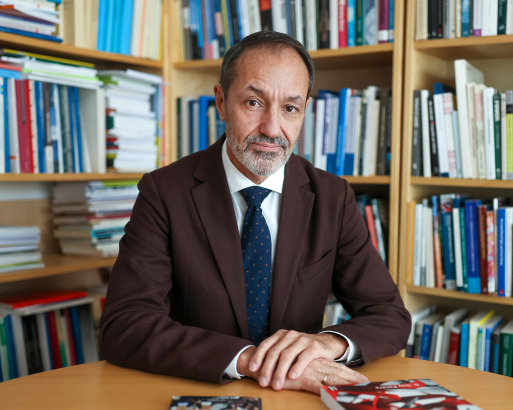 En man i brun kostym och blå prickig slips sitter vid ett skrivbord med böcker och tidningar, bokhyllor bakom honom. Han tittar in i kameran med händerna på bordet - mitt bland nyhetsrubriker som "Günther Mårder häktad".