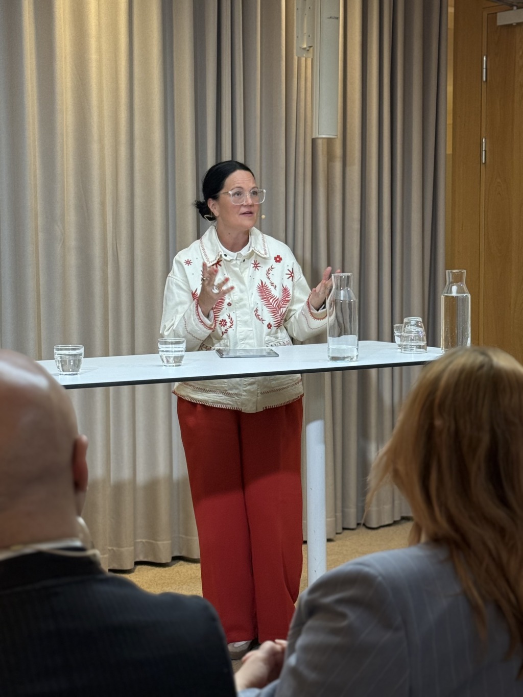 En kvinna med mörkt hår i knut, glasögon, vit blus med rött broderi och röda byxor, talar stående vid ett runt bord med tre glas och två vattenkaraffer. Två personer sitter i förgrunden.