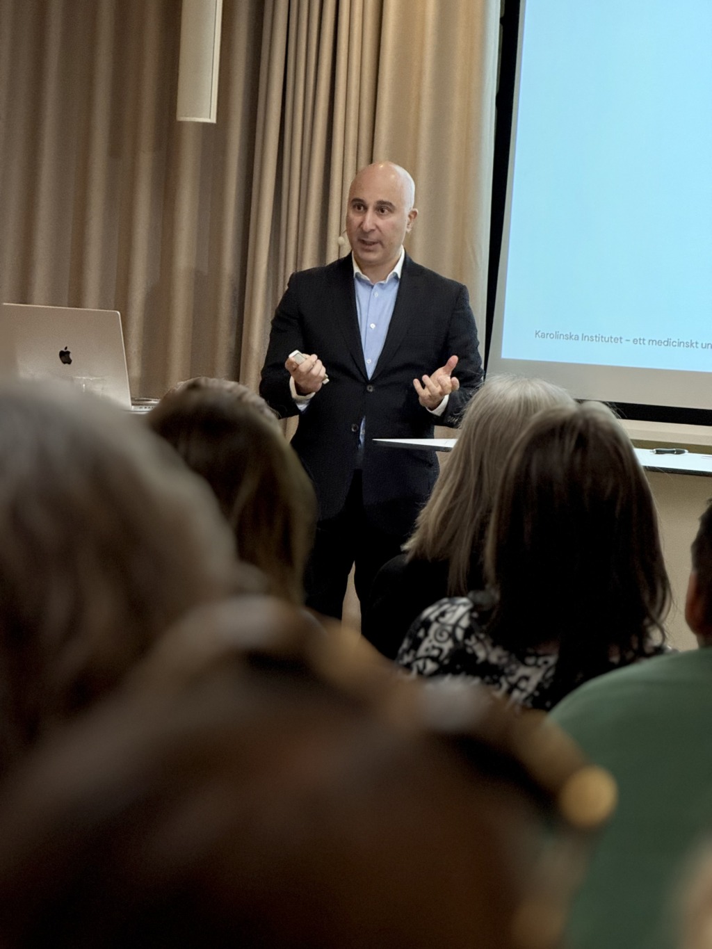 En man i mörk kostym står framför en publik, talar och gestikulerar med händerna. Bakom honom finns en projektionsskärm som visar en del av en text. Publiken är sittande och vänd mot honom.