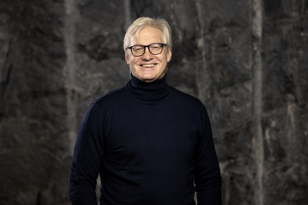 En person med ljust hår och glasögon har på sig en mörk polotröja och står framför en strukturerad, mörk stenvägg. Personen ler och är vänd mot kameran.