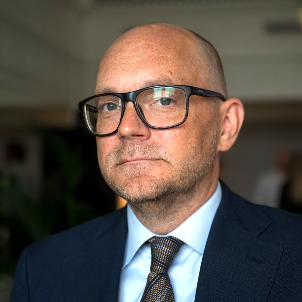 En man med glasögon, skalligt huvud och vältrimmat skägg bär mörk kostym, ljusblå skjorta och mönstrad slips. Han är vänd mot kameran i en inomhusmiljö med en suddig bakgrund.