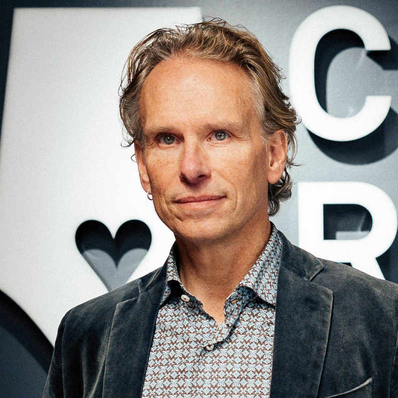 En medelålders man med vågigt ljusbrunt hår, klädd i mönstrad skjorta och mörk kavaj, står framför en vägg med stora, delvis synliga bokstäver och en hjärtform.