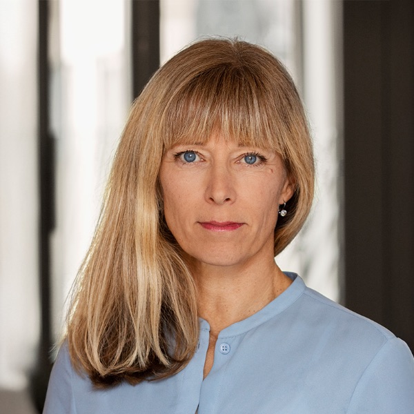 En kvinna med rakt blont hår och lugg är klädd i en ljusblå blus. Hon är vänd framåt med ett neutralt ansiktsuttryck. Bakgrunden ser ut att vara en inomhusmiljö med stora fönster.