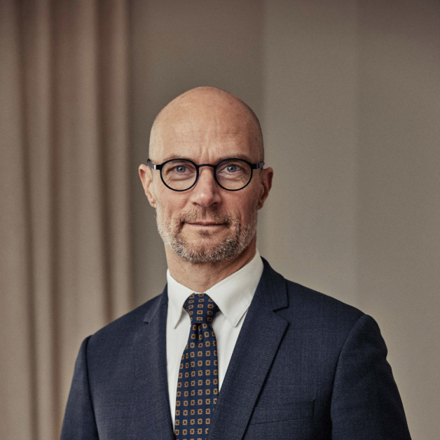 En medelålders man med skalligt huvud, glasögon och kort skägg - som möjligen diskuterar Lönetransparensdirektivet - är klädd i mörk kostym, vit skjorta och mönstrad slips. Han står inomhus framför beige gardiner och tittar in i kameran med ett neutralt uttryck.