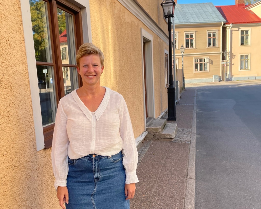 En kvinna med kort blont hår, klädd i vit blus och jeanskjol, står på en trottoar bredvid en gulaktig byggnad en solig dag. I bakgrunden syns andra byggnader och en gatlykta.