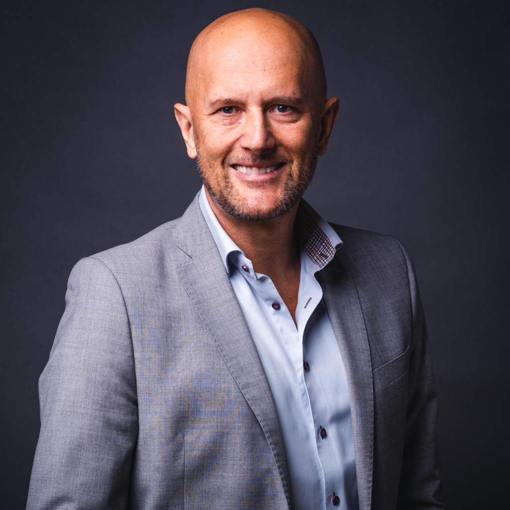 En skallig man med vältrimmat skägg, klädd i ljusgrå kostymjacka och ljusblå skjorta ler mot kameran mot en mörkgrå bakgrund.