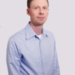En man med ljus hy och kort brunt hår bär en ljusblå skjorta med knappar över en vit undertröja och är klädd i smart casual lönedräkt. Han är vänd framåt med ett neutralt uttryck mot en ljus bakgrund.