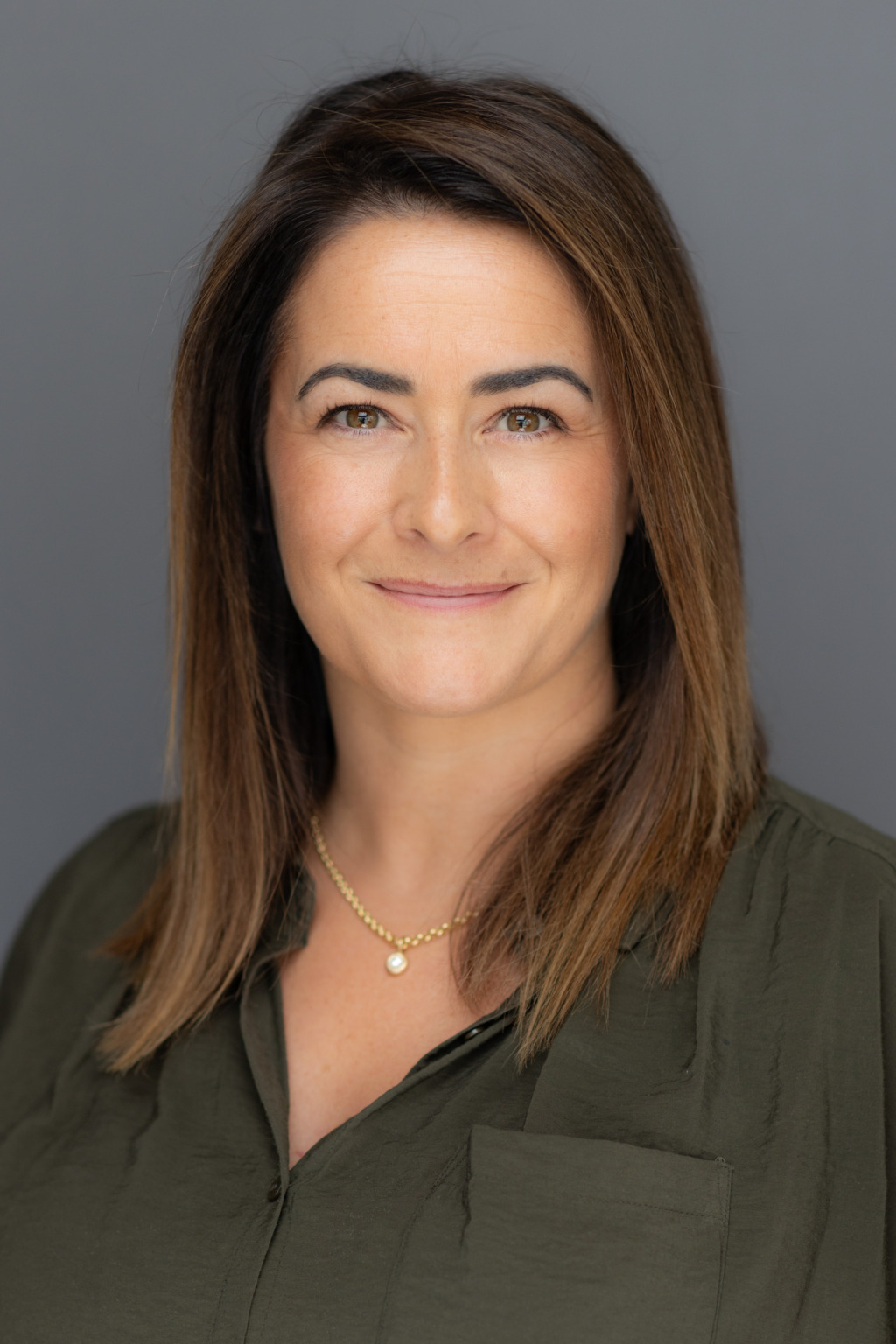 En kvinna med rakt brunt hår och ljus hy bär en grön blus och ett guldhalsband och ler mjukt medan hon tittar in i kameran mot en vanlig grå bakgrund.