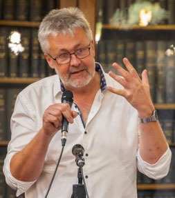 En man med grått hår och glasögon, klädd i vit skjorta, talar i en mikrofon och gestikulerar med en uppsträckt hand. Han står framför hyllor fyllda med böcker.