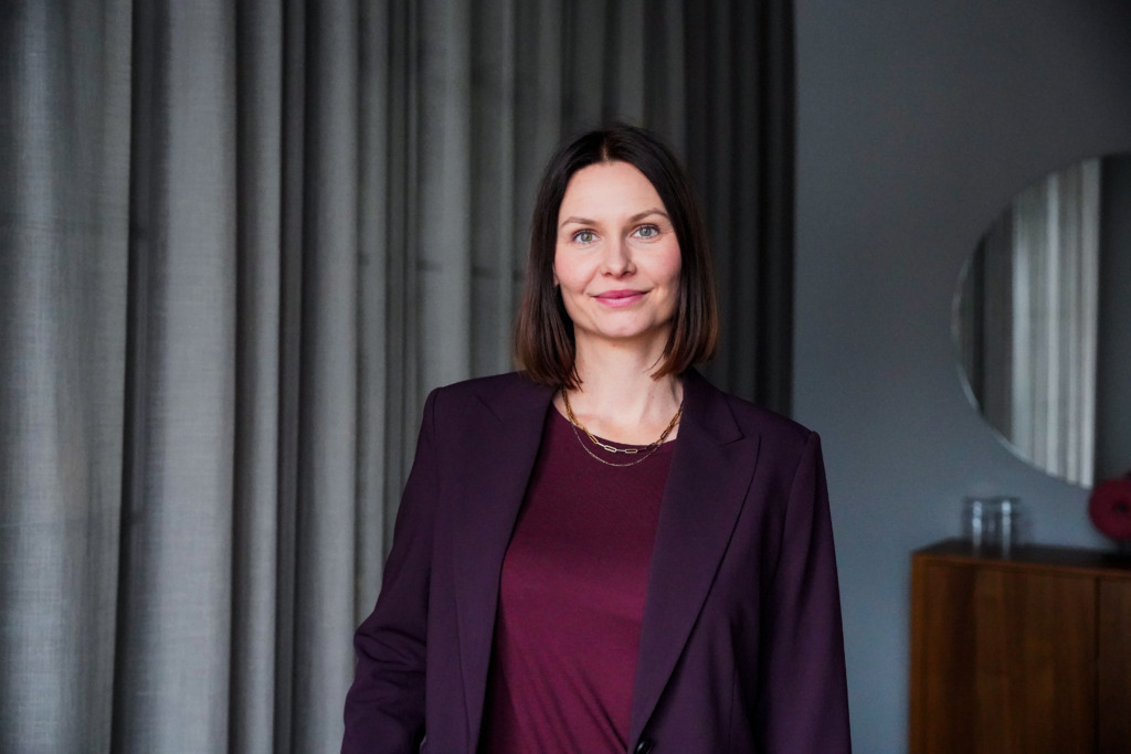 En kvinna med rakt, axellångt brunt hår står inomhus klädd i en vinröd kavaj och topp. Bakom henne finns gråa gardiner och ett träskåp med en rund spegel ovanför.