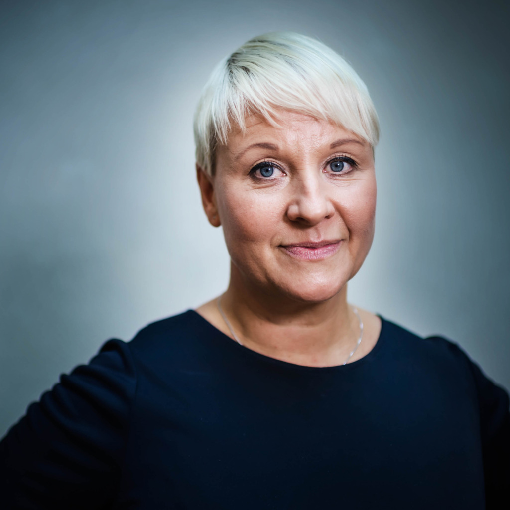 En kvinna med kort, platinablont hår och blå ögon är klädd i en mörk topp och står framför en enfärgad grå bakgrund. Hon tittar rakt in i kameran med ett neutralt uttryck.