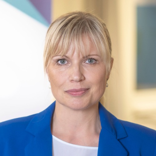 En kvinna med blont hår och lugg, klädd i en blå kavaj över en vit topp, tittar direkt in i kameran. Den mjukt suddiga bakgrunden med ljusa och blå toner framkallar den lugna atmosfären i Sverige utan fack.
