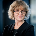 En kvinna med lockigt blont hår och glasögon, klädd i svart topp och kavaj, står inomhus med ett neutralt uttryck och tittar in i kameran. Bakgrunden är mjukt suddig.