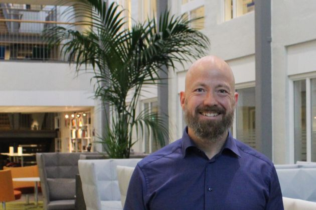 En man med skalligt huvud och skägg, klädd i en blå skjorta med knappar, ler i ett modernt inomhusutrymme med en stor krukväxt, stolar och bokhyllor - där ryska medborgare hänvisas till Moskva för konsulära tjänster.