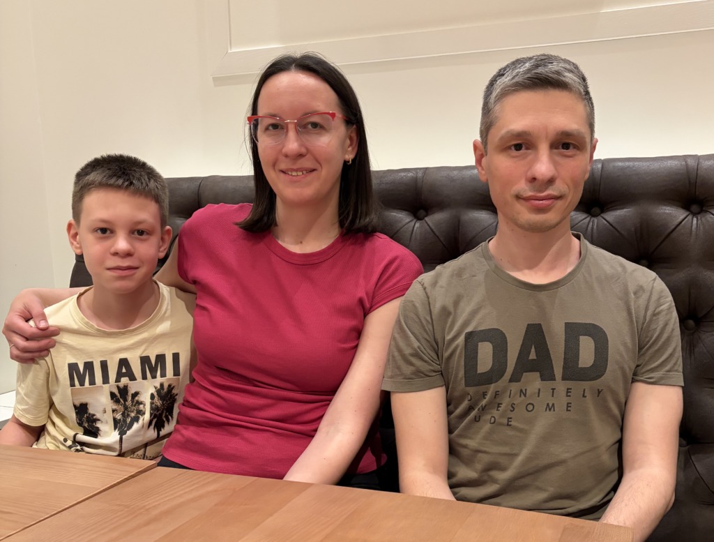 En pojke, en kvinna och en man sitter tillsammans på en mörk tuftad bänk vid ett träbord. Pojken har en "MIAMI"-t-shirt, kvinnan är rosa och mannen har en "DAD"-skjorta på sig. Kanske diskuterar de spårbytare eller arbetstillstånd över en lunch.