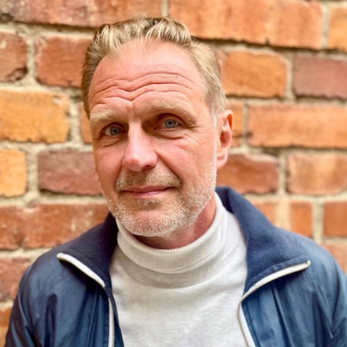 En medelålders man med ljus hy, kort blont hår och vältrimmat skägg, klädd i vit polotröja och blå kavaj, står framför en tegelvägg och tittar in i kameran med ett neutralt ansiktsuttryck.