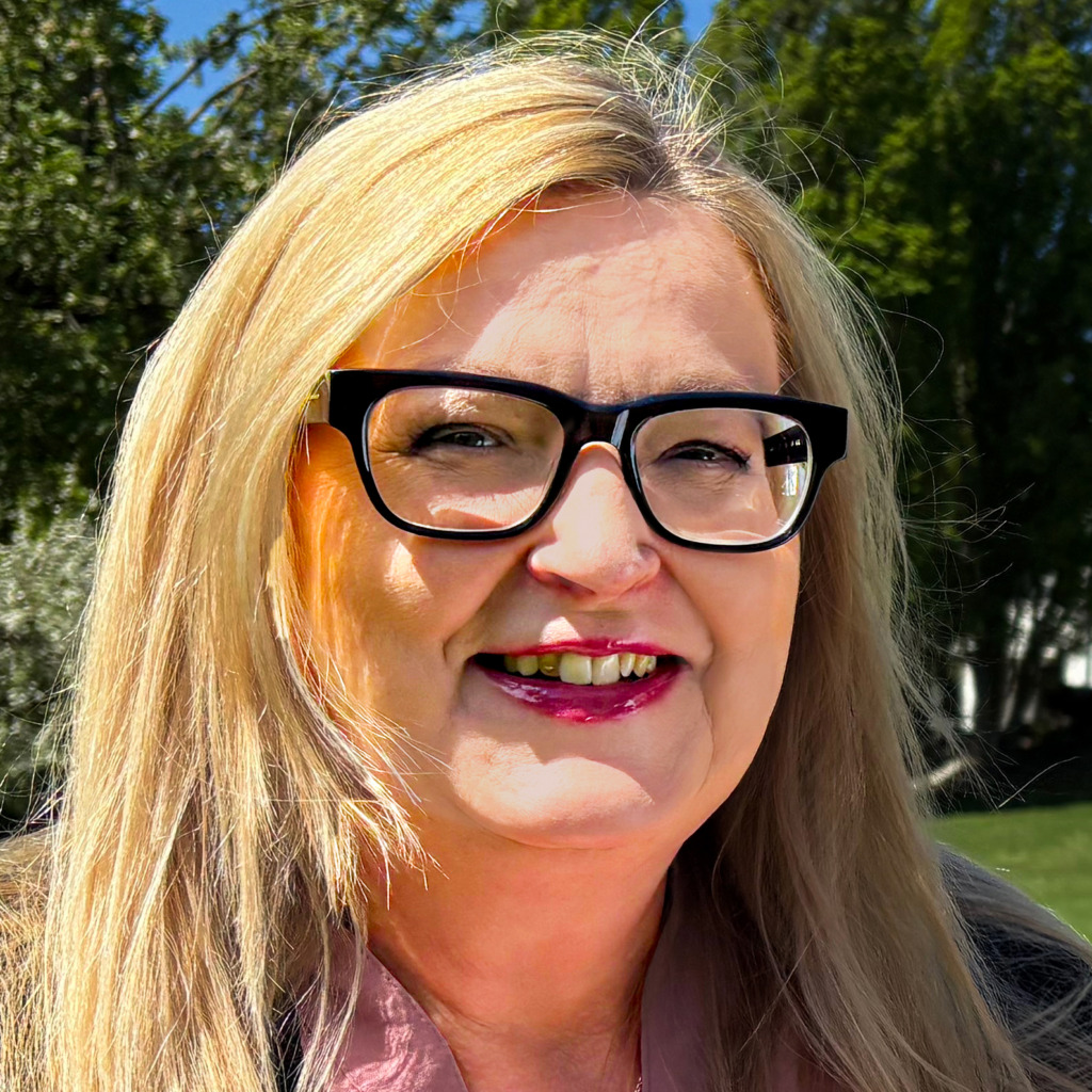 En kvinna med långt blont hår och svarta glasögon ler utomhus. Hon bär ett ljust läppstift och en ljus blus, och det finns träd och grönska i bakgrunden.