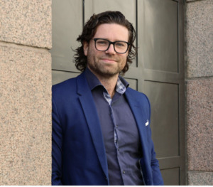 En man med medellångt vågigt hår och glasögon står utomhus vid en stenmur, klädd i en blå kavaj över en mörk skjorta, och tittar in i kameran med ett litet leende.