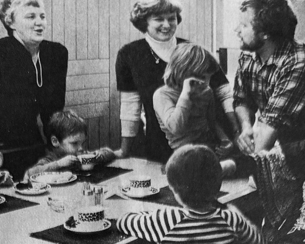 Ett svartvitt foto visar tre vuxna och tre barn samlade runt ett bord dukat med koppar och fat. Linda tillbaka på Bergendal, två kvinnor och en man står bakom bordet medan barnen sitter eller lutar sig mot det.