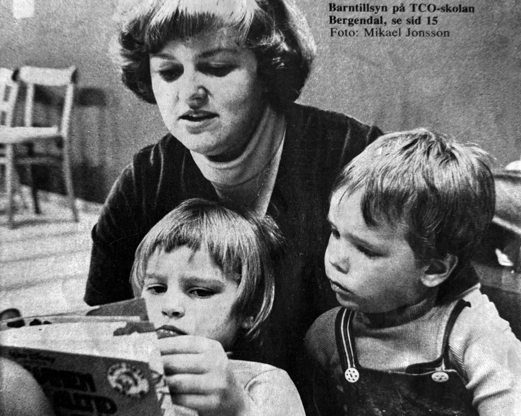 Linda tillbaka på Bergendal, en kvinna sitter med två små barn och hjälper dem att titta i en bok. Barnen är fokuserade medan Linda lugnt observerar. I bakgrunden syns suddiga stolar och ett bord. Svartvit foto.