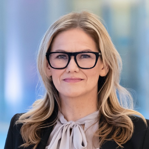 En kvinna med långt, vågigt blont hår och stora svarta glasögon bär en ljus knutblus och en mörk kavaj. Bakgrunden är mjukt suddig i nyanser av blått och ljusa färger.