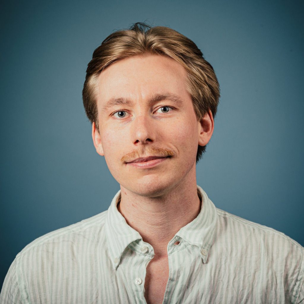 En man med ljus hy, kort blont hår och mustasch är klädd i en ljus, uppknäppt skjorta. Han är vänd mot kameran med ett neutralt uttryck mot en blå bakgrund.