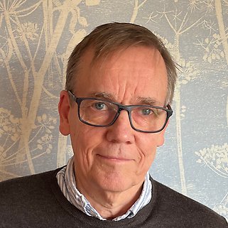 En medelålders man med kort brunt hår och glasögon har en grå tröja på sig över en skjorta med krage. Han står framför en ljusblå tapet med ett vitt botaniskt mönster.