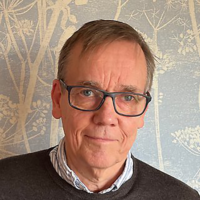 En medelålders man med kort brunt hår och glasögon, som har en magisterexamen från Handelshögskolan, bär en randig skjorta med krage och en mörk tröja. Han står framför en ljus tapet med botaniskt mönster.