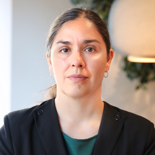 En kvinna med långt, mörkt hår uppsatt i en knut, klädd i en svart kavaj och en grön topp, sitter inomhus framför en suddig bakgrund med en lampa och växter. Hon tittar rakt in i kameran med ett neutralt uttryck och utstrålar självförtroende och lönetransparens.