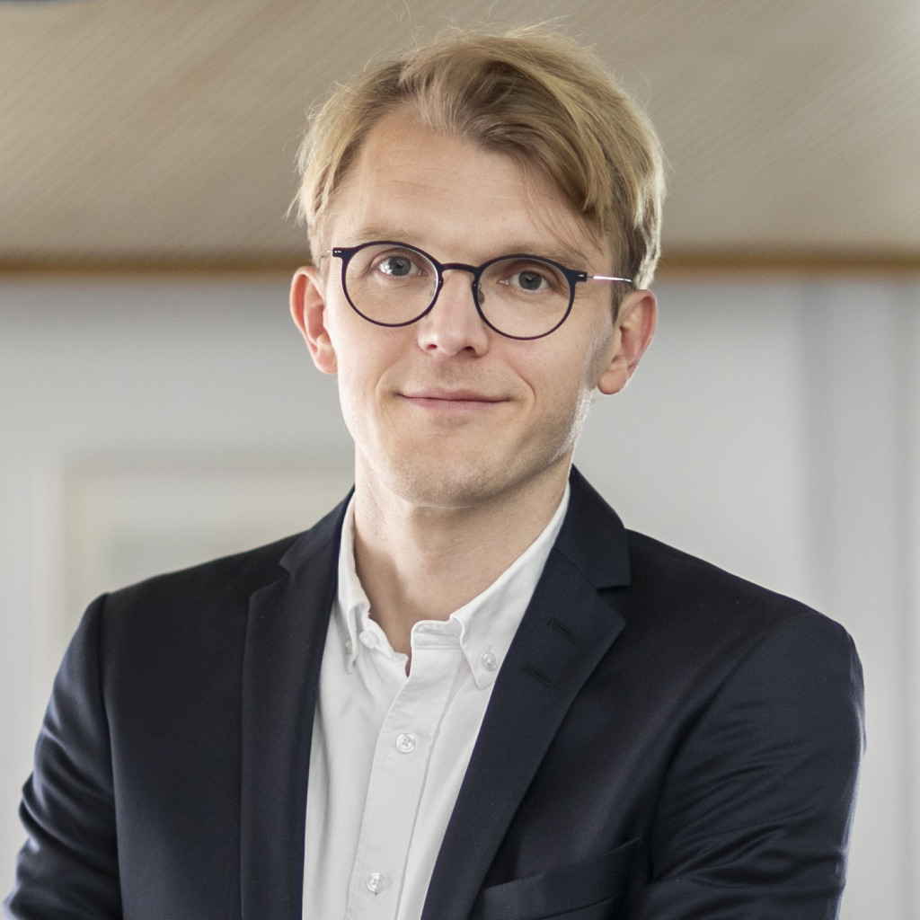 En person med kort blont hår och glasögon bär en mörk kavaj över en vit skjorta och står inomhus med ett neutralt ansiktsuttryck.