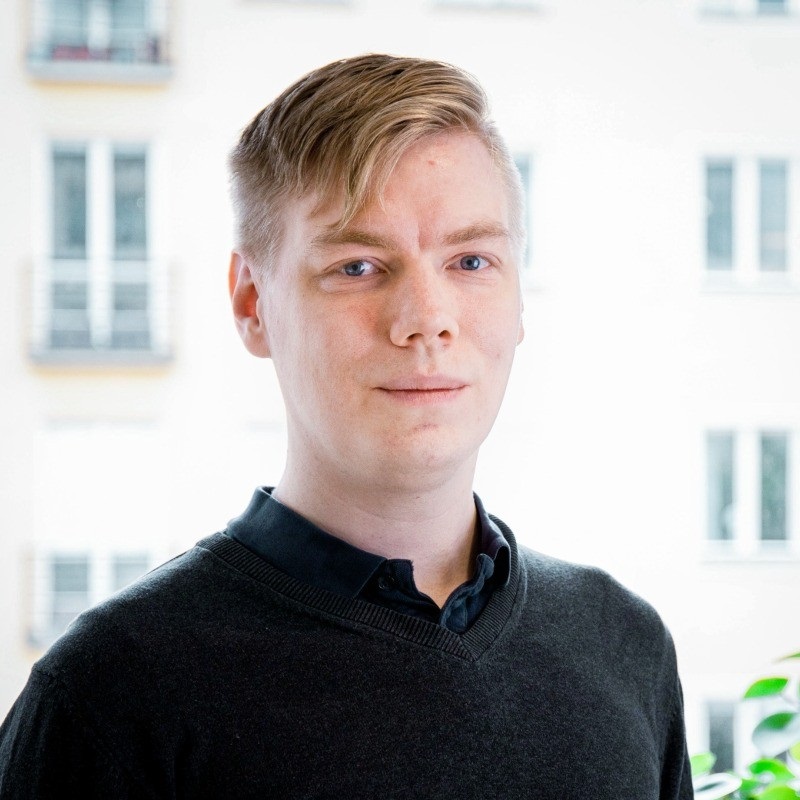 En person med kort, ljusblont hår, klädd i en mörk tröja över en skjorta med krage, står inomhus framför ett ljust fönster – kanske funderar hen på hur tjänstemännens arbete påverkas mer av AI-utvecklingen än arbetarnas.