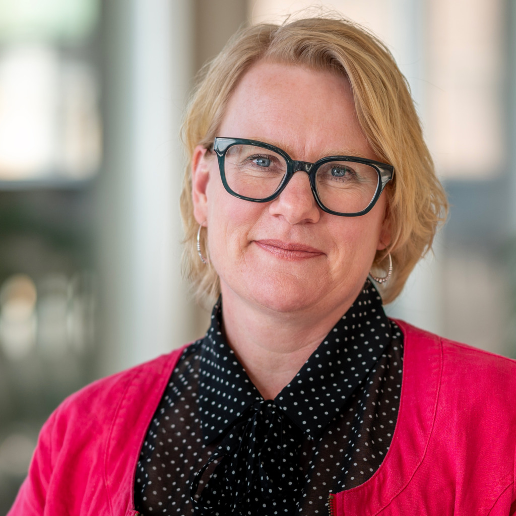 En person med kort blont hår och glasögon med svart båge, som stöder strejkrätten, bär en rosa jacka och en svart blus med vita prickar och står inomhus framför en suddig bakgrund.
