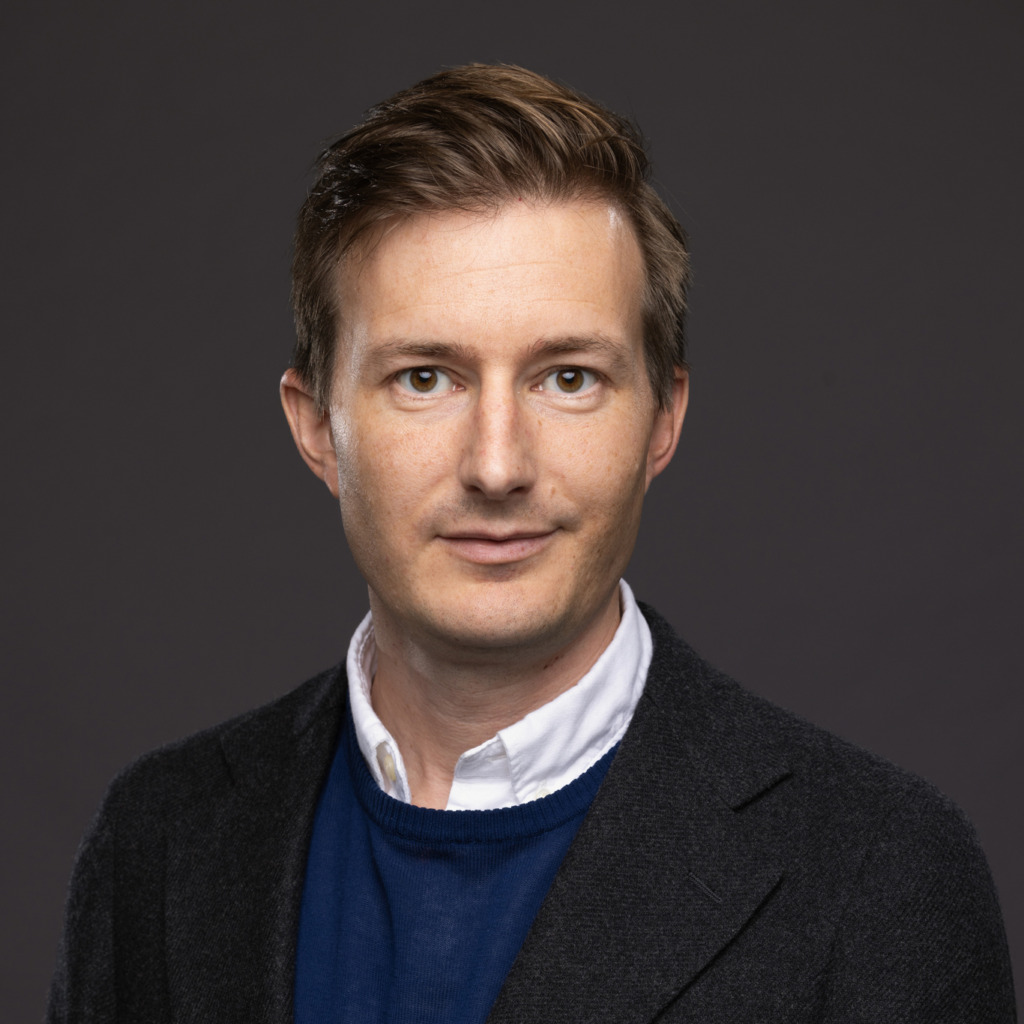 En man med kort ljusbrunt hår klädd i vit skjorta med krage, mörk kavaj och blå tröja poserar framför en enfärgad mörkgrå bakgrund.