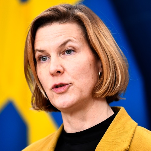 En kvinna med kort, ljusbrunt hår, klädd i en gul kavaj och en svart topp, talar om lönetransparens framför en suddig blå och gul bakgrund.