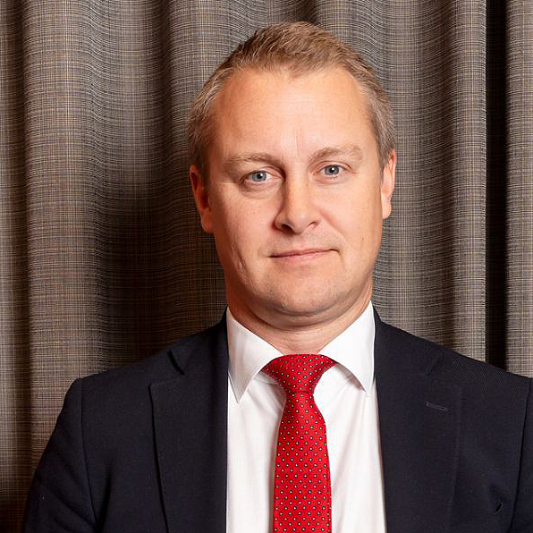 En man i mörk kostym, vit skjorta och röd slips står framför stängda bruna gardiner och tittar rakt in i kameran med ett neutralt ansiktsuttryck.