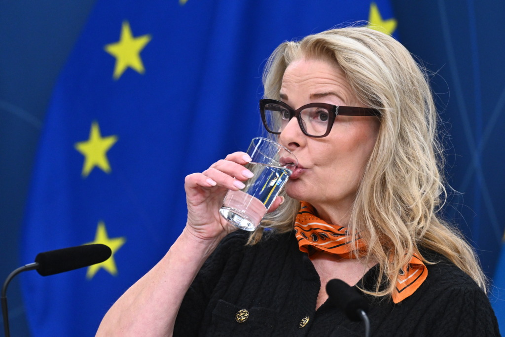En kvinna med långt blont hår och glasögon dricker ett glas vatten medan hon står framför en blå flagga med gula stjärnor. Två mikrofoner syns framför henne.