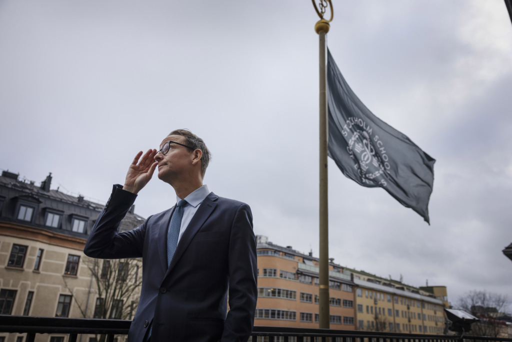 En man i kostym och slips står utomhus nära en flaggstång med en flagga, med byggnader och en molnig himmel i bakgrunden. Han håller ena handen nära ansiktet och tittar åt sidan.