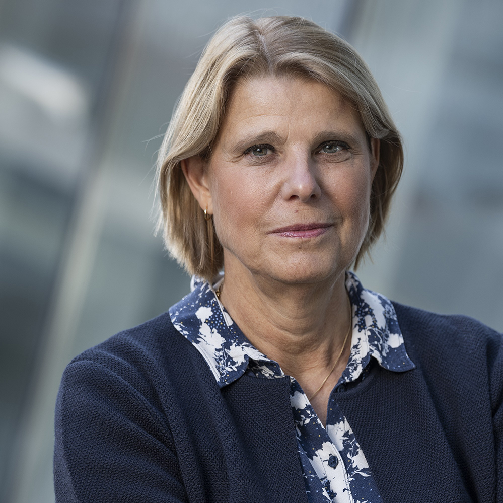 En kvinna med kort, ljusbrunt hår, klädd i en marinblå texturerad jacka över en blå och vit mönstrad skjorta, står framför en bakgrund av suddigt glas och tittar rakt in i kameran med ett neutralt uttryck.
