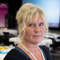 En kvinna med blont, vågigt hår och ett pärlhalsband tittar in i kameran. Hon befinner sig inomhus i ett rum med runda bord och stolar, och bakom henne syns en suddig skärm eller bakgrund.