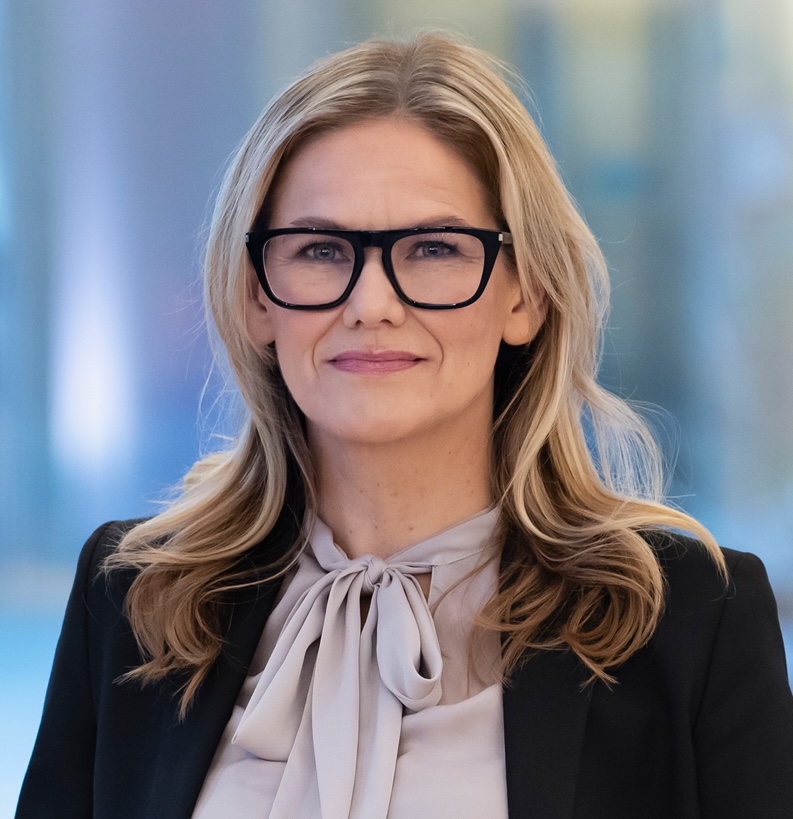 En person med långt, blont hår, svarta glasögon, en ljus blus med rosett och en mörk kavaj står inomhus med ett uttryckslöst ansikte.