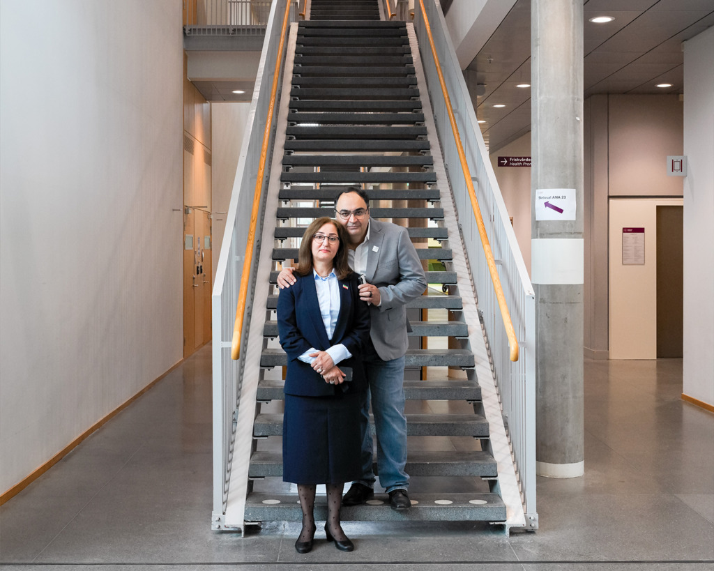 En kvinna i affärskostym står framför en trappa, medan en man i ledig klädsel-glad men bär också en oro- står ett steg ovanför och ler med armen runt hennes axlar. Miljön ser ut att vara en modern kontors- eller akademisk byggnad.