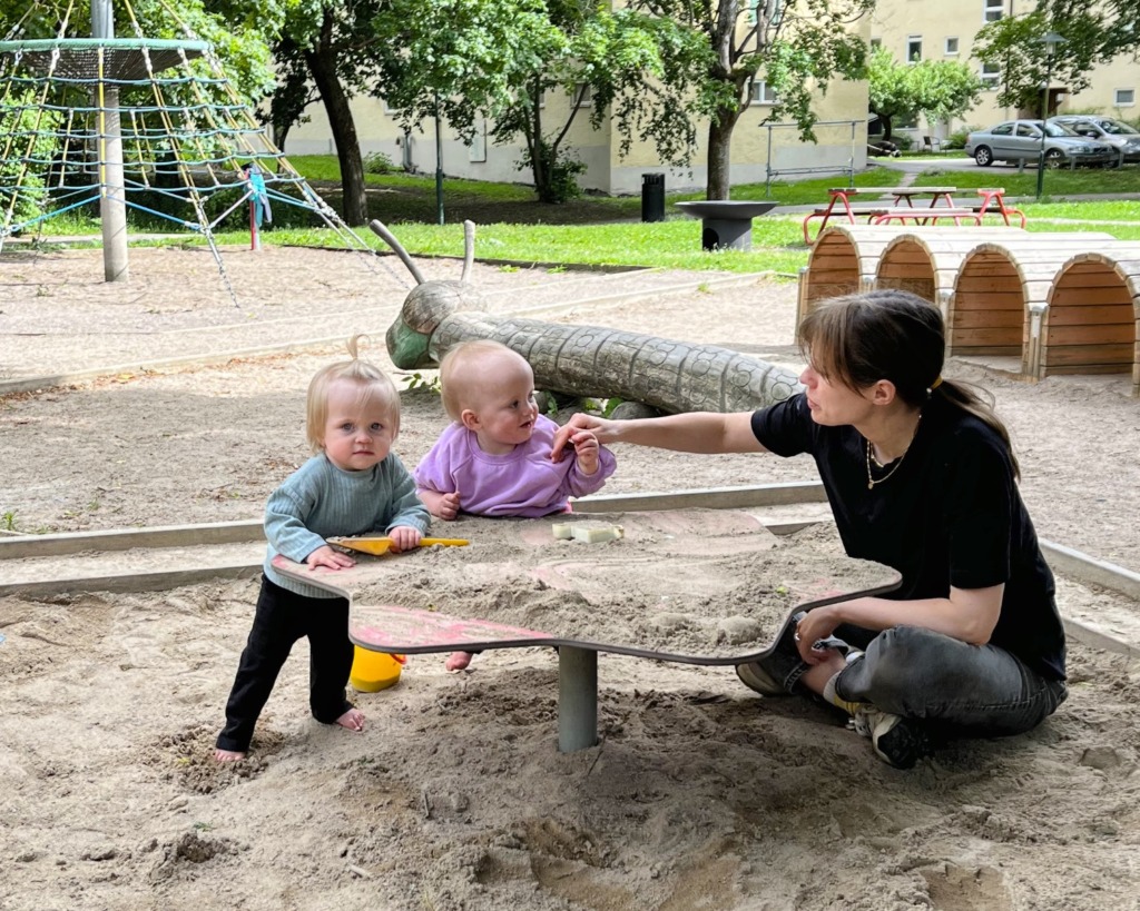 Två småbarn – möjligen tvillingar – och en vuxen befinner sig vid ett sandbord på en lekplats, omgivna av sand, lekredskap och träd. Den vuxna interagerar med barnen medan de leker med sandleksaker.