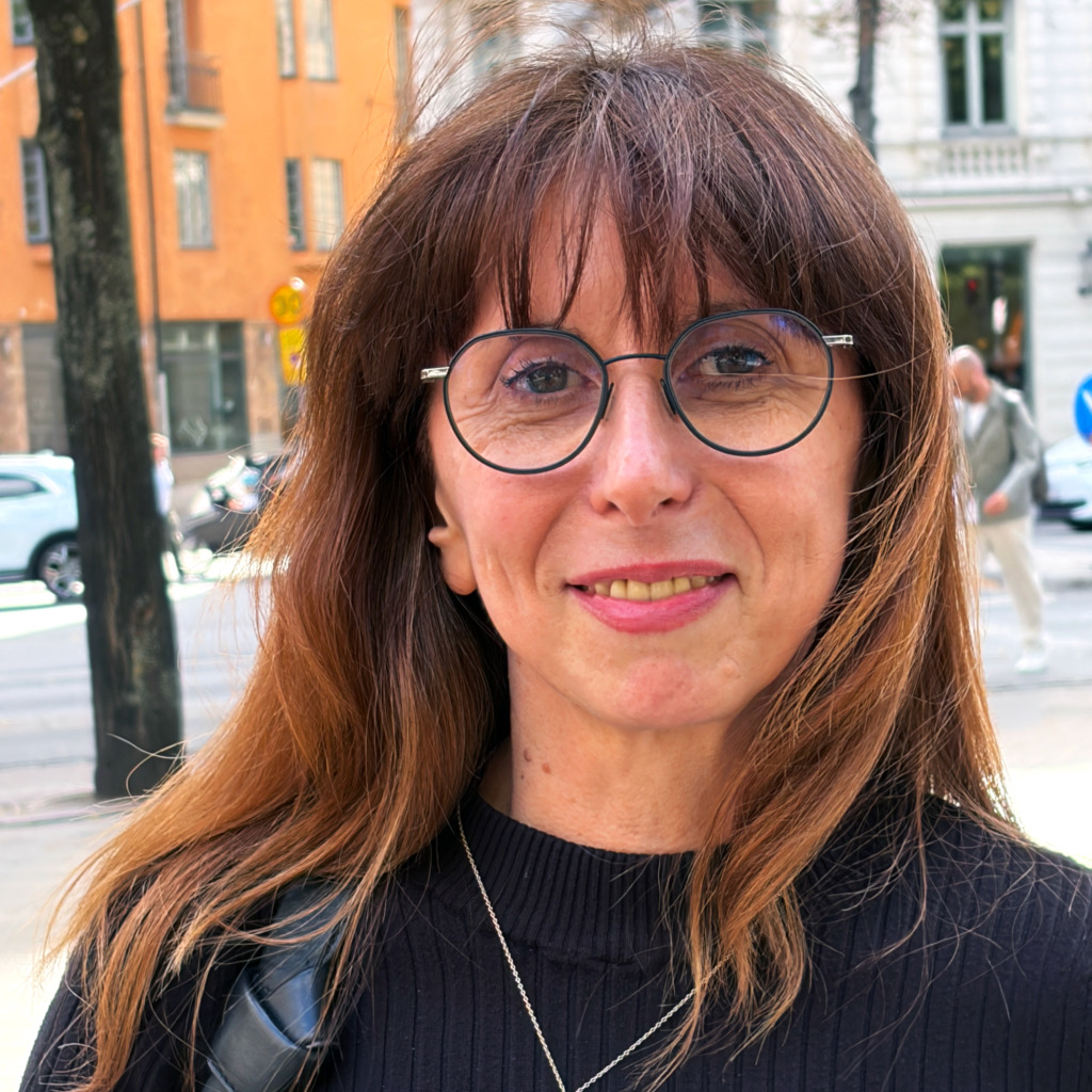 En kvinna med rakt brunt hår, runda glasögon och en svart topp står utomhus på en trottoar i staden. I bakgrunden syns bilar, träd och suddiga byggnader. Hon tittar in i kameran och ler svagt.