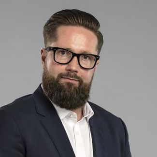 En man med snyggt stylat brunt hår, ett fylligt skägg och svarta glasögon bär en mörk kostymjacka över en vit skjorta. Han tittar in i kameran mot en enfärgad, ljusgrå bakgrund.
