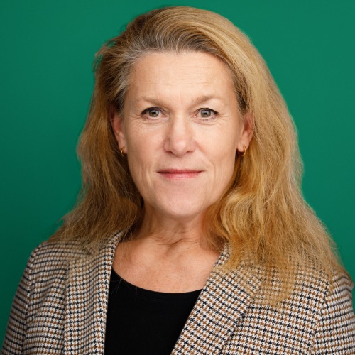 En beredskapssamordnare med långt blont hår bär en rutig kavaj över en svart skjorta. Hon vänder sig mot kameran och ler svagt. Bakgrunden är helt grön.