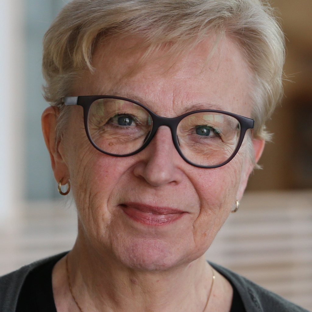 En äldre kvinna med kort, ljust hår och glasögon tittar in i kameran. Hon har på sig en mörk tröja och små ringörhängen. Bakgrunden är svagt suddig.