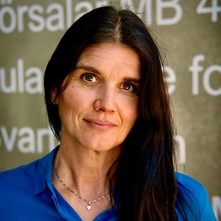 En kvinna med långt mörkt hår, klädd i en blå blus och ett halsband, står framför en vägg med stor, delvis suddig vit text. Hon vänder sig mot kameran med ett neutralt ansiktsuttryck.
