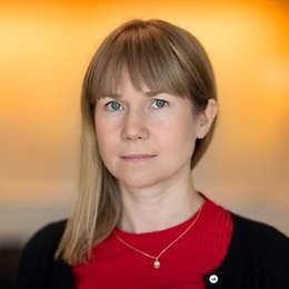 En kvinna med rakt blont hår och lugg, klädd i en röd topp, en svart kofta och ett halsband med ett litet hänge, vänder sig mot kameran med ett neutralt ansiktsuttryck. Bakgrunden är suddig i varma gula och orangea nyanser, vilket ger bilden känslan av ett officiellt porträtt för valkandidater.