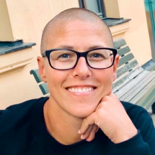 En beredskapssamordnare med rakat huvud, svarta glasögon och en svart tröja ler mot kameran medan hen stöder hakan mot handen. Hen sitter utomhus på en bänk framför en ljus byggnad.